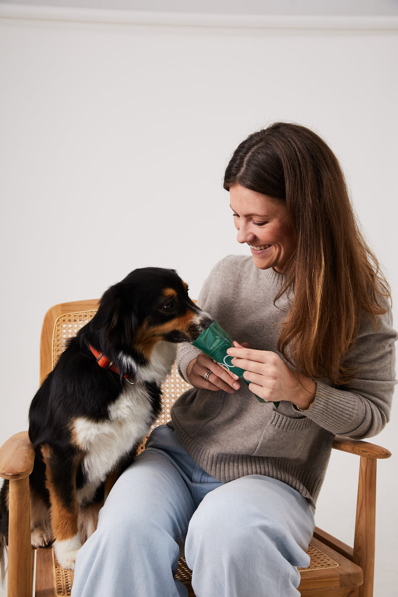 Frau und Hund sitzen auf Stuhl. Die Frau hält dem Hund einen DOG1 Beutel hin und der Hund steckt die Schnauze rein.